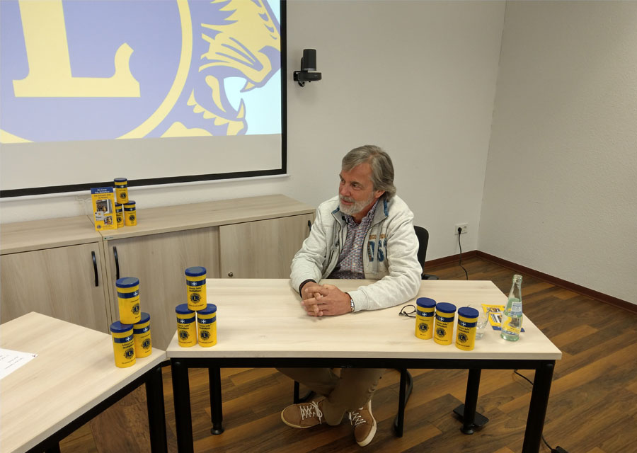 Werner Burg am Tisch im Gespräch mit TACHELES, um ihn herum mehrfach gestapelt die gelbe Notfall-Box, im Hintergrund das Logo des Lion´s Clubs an der Wand