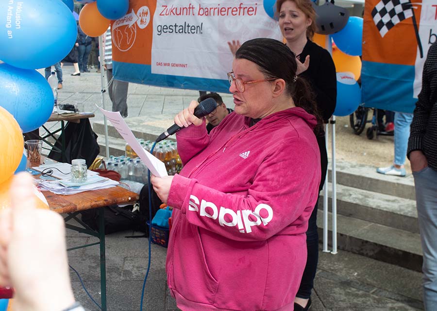 TACHELES-Redakteurin Bea hält eine Rede mit Mikrofon in der Hand, vor und hinter ihr Banner und Luftballons in Blau und Orange