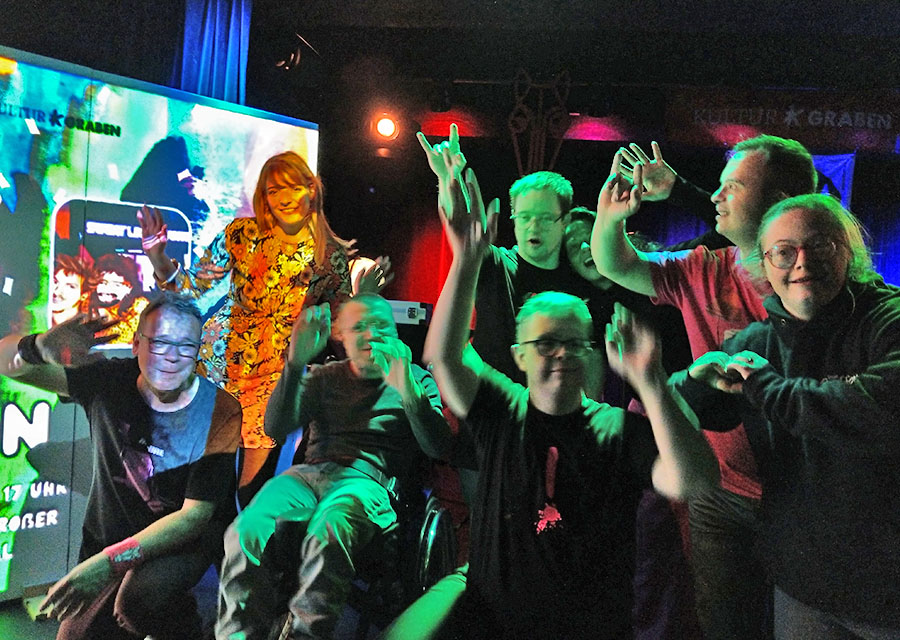 TACHELES-Patrick jubelt mit Musikern in die Kamera, dunkle, bunte Partybeleuchtun, links eine bunte Leinwand.