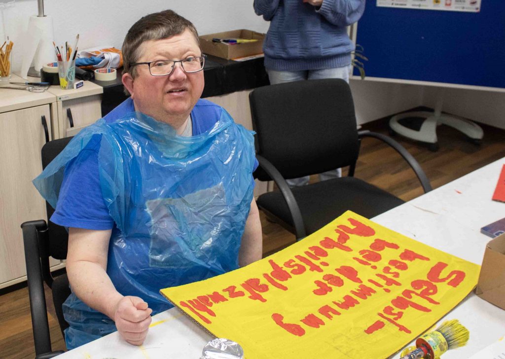 Eine Person sitzt vor einem gemalten Schild in gelb und rot mit den Worten: Selbstbestimmung bei der Arbeitsplatzwahl