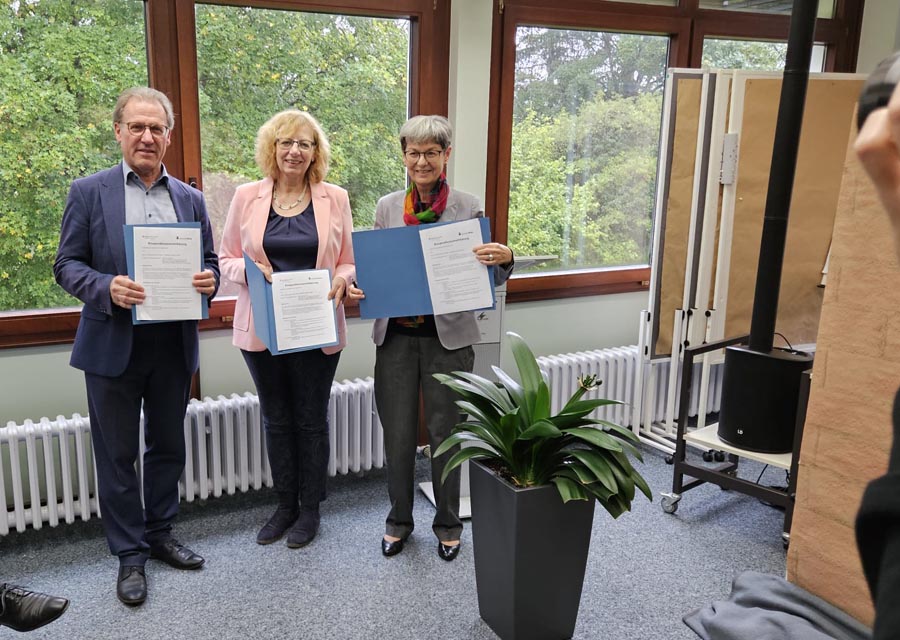 3 Menschen stehen in vor Fenstern mit Blick ins Grüne und halten blaue Mappen mit unterzeichneten Verträgen in die Kamera.