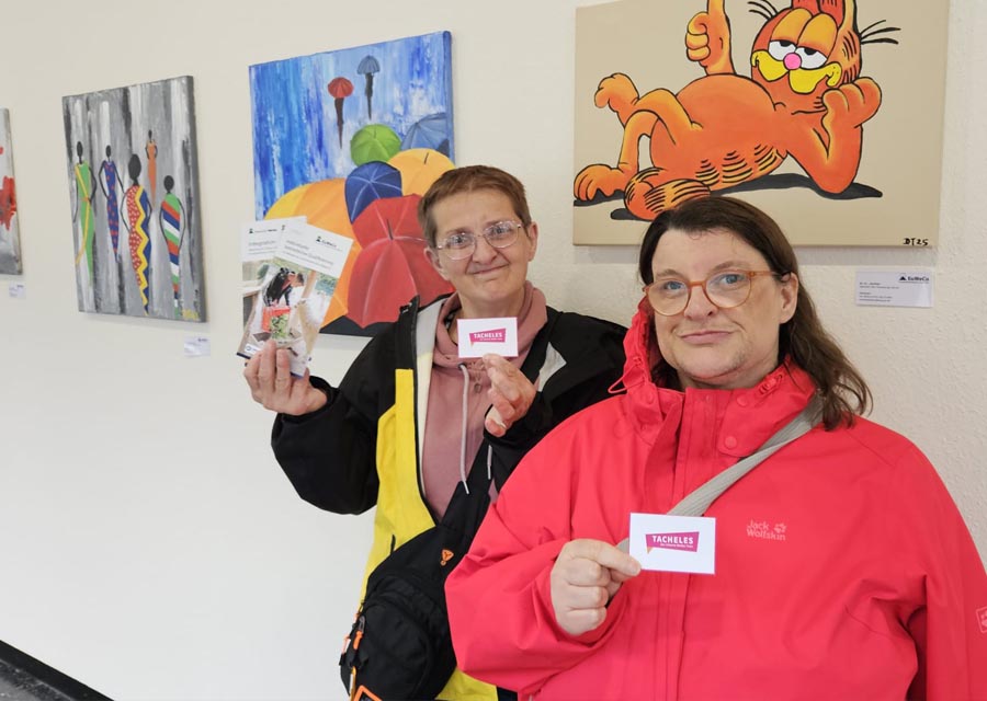 Bea und Martina von TACHELES stehen in einem Flur vor ganz verschiedenen Bildern an der Wand und halten das TACHELES-Logo und Euweco-Flyer hoch