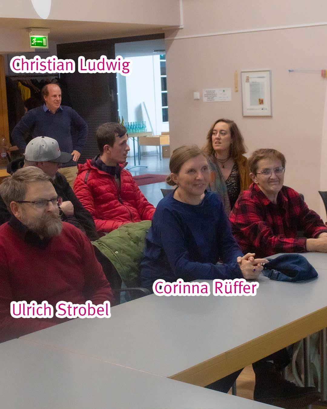 Eine Gruppe von Leuten sitzt auf Stühlen hinter Tischen und guckt an der Kamera vorbei nach vorne.