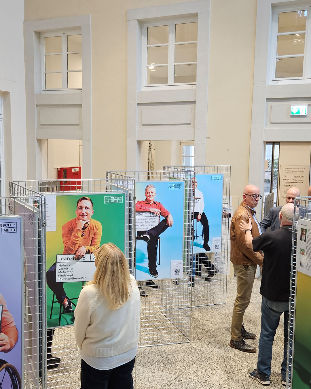 Blick von oben auf die Ausstellung: Foyer eines großen Hauses, helle Wände und Boden, Drahtaufsteller in Zickzackform aufgestellt, daran hängen Bilder von Leuten mit Beschriftung.
