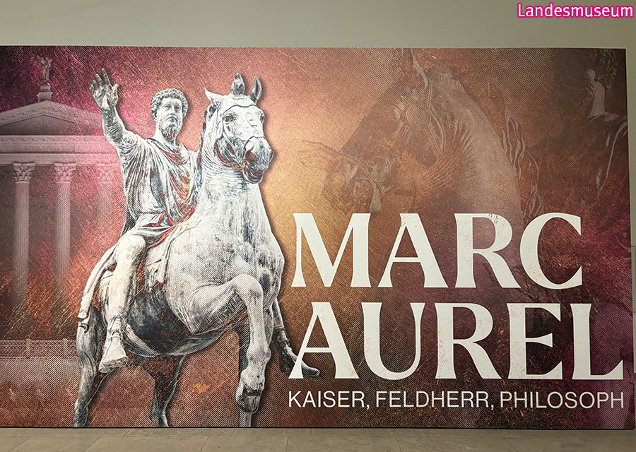 Ein großes Plakat mit Marc Aurel in grau auf einem Pferd und rechts sein Name