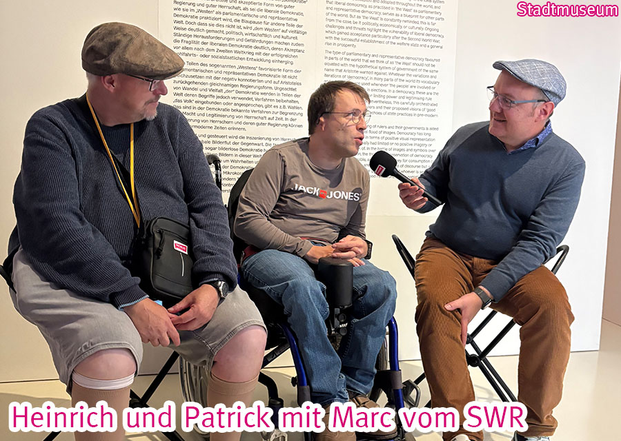Heinrich, Patrick (TACHELES) und Marc vom SWR sitzen mit Mikro vor einer beschrifteten Wand