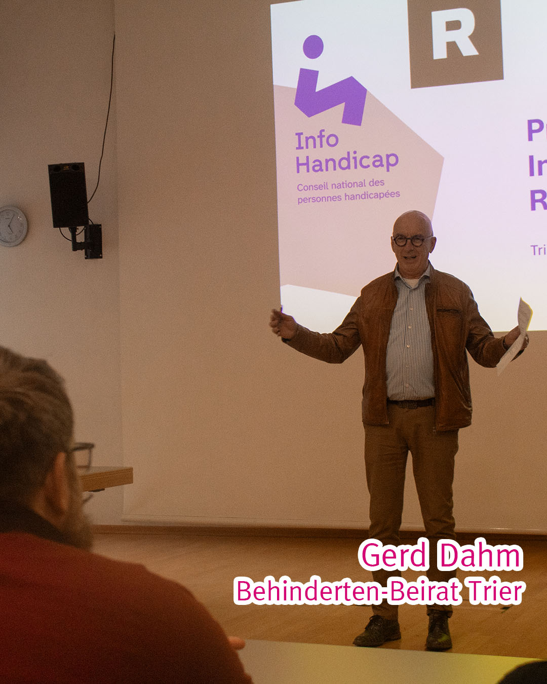 Gerd Dahm vor einer Leinwand, gesehen über die Schulter eines Rollstuhlfahrers.