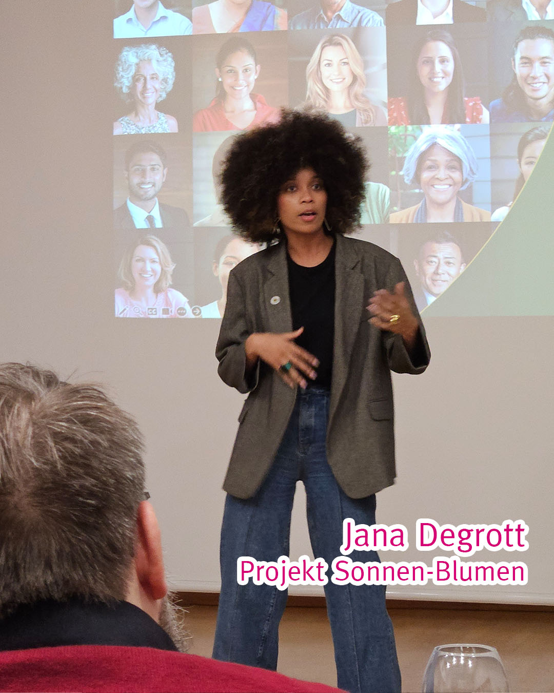 Jana Degrott mit Afro vor einer Leinwand, gesehen über die Schulter eines Rollstuhlfahrers.