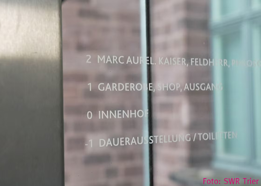 Weiße Schrift auf Glas, schwer zu lesen, im Hintergrund verschwommen eine Stein-Mauer
