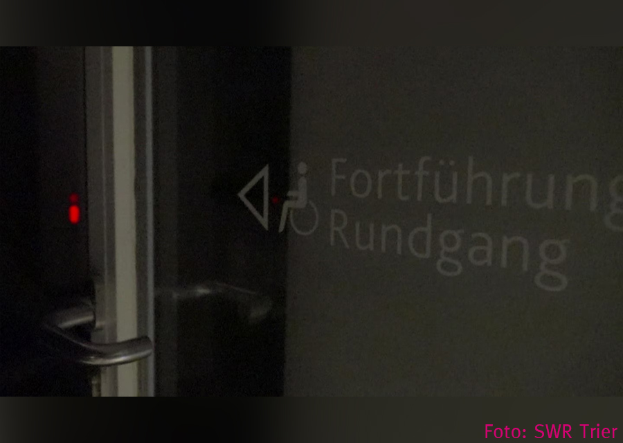 sehr dunkel, Schrift: Fortführung Rundgang mit Piktogrammen Rollstuhl und Pfeil nach links, dort ein kleines rotes Licht in der Dunkelheit