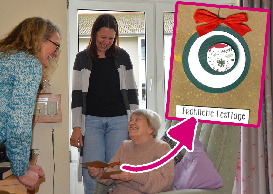 Drei lachende Frauen, zwei stehend, eine ältere sitzend mit einer Weihnachtskarte in der Hand, alle lachen. Die Karte rechts oben noch mal groß im Bild. Ein Pfeil zeigt darauf. Alle freuen sich über die Karte, die sie von TACHELES bekommen haben.