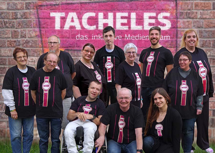 12 Menschen, alle im schwarzen TACHELES-T-Shirt, stehen auf einer Wiese vor einer Steinwand mit TACHELES-Logo-Grafitti