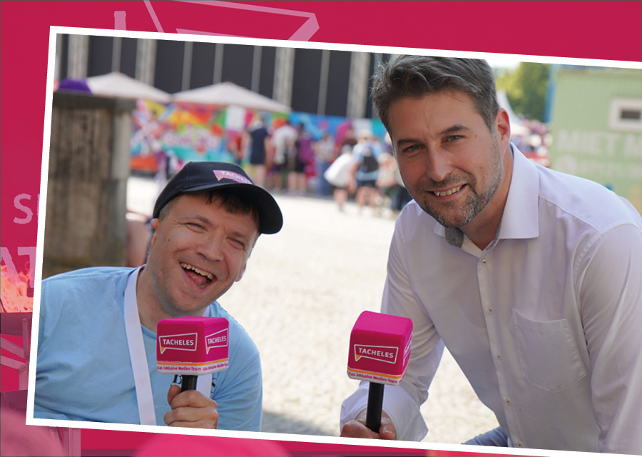 Patrick von TACHELES spricht mit Herrn Conradt, dem Oberbürgermeister von Saarbrücken. Beide halten pinke TACHELES-Mikrofone in der Hand und lächeln in die Kamera. Im Hintergrund sind unscharf bunte Stände und Menschen zu sehen.