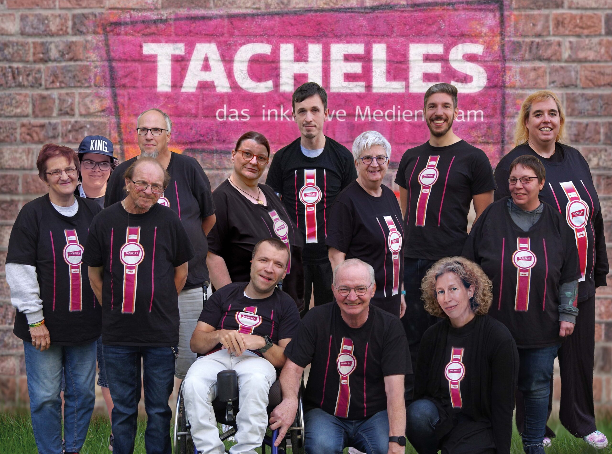 Gruppenbild von TACHELES. Die Redaktion steht gemeinsam vor einer Wand mit dem TACHELES-Logo in Grafitti-Optik. Alle tragen das schwarze T-Shirt mit TACHELES-Logo und schauen in die Kamera.